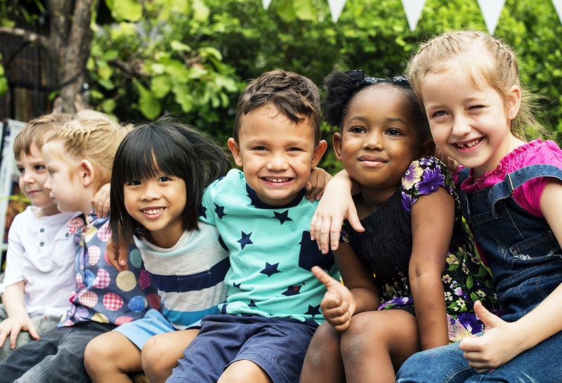 Five children sitting together outdoors, smiling and embracing with green foliage in background.